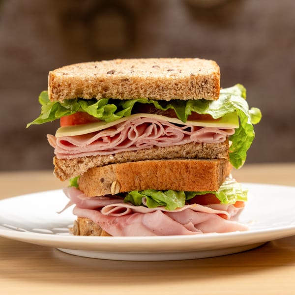 A tall, stacked sandwich with whole wheat bread, ham, cheese, lettuce, and tomato on a white plate.