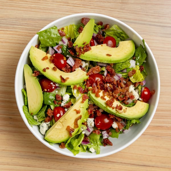 Overhead view of a salad with lettuce, avocado, cherry tomatoes, bacon bits, red onion, and feta cheese.