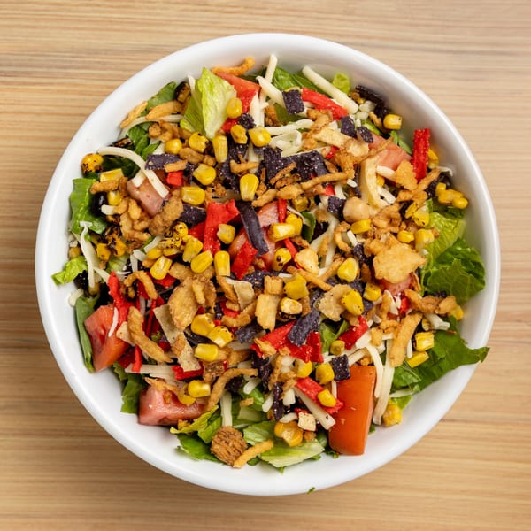 A vibrant salad in a white bowl, topped with corn, black beans, cheese, and crispy tortilla strips.