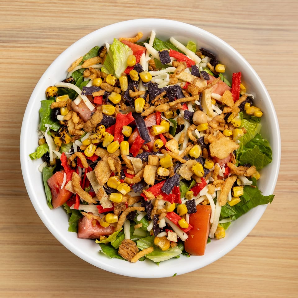 A vibrant salad in a white bowl, topped with corn, black beans, cheese, and crispy tortilla strips.