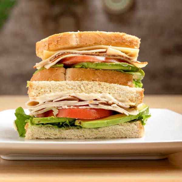 A towering club sandwich with turkey, cheese, lettuce, tomato, and avocado on a white plate.