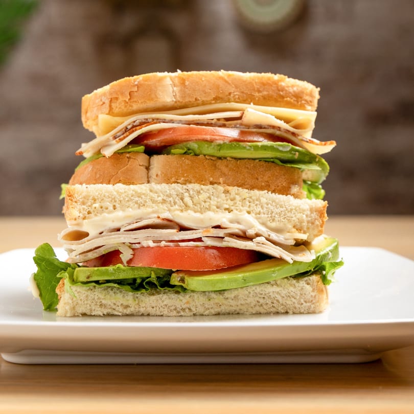 A towering club sandwich with turkey, cheese, lettuce, tomato, and avocado on a white plate.
