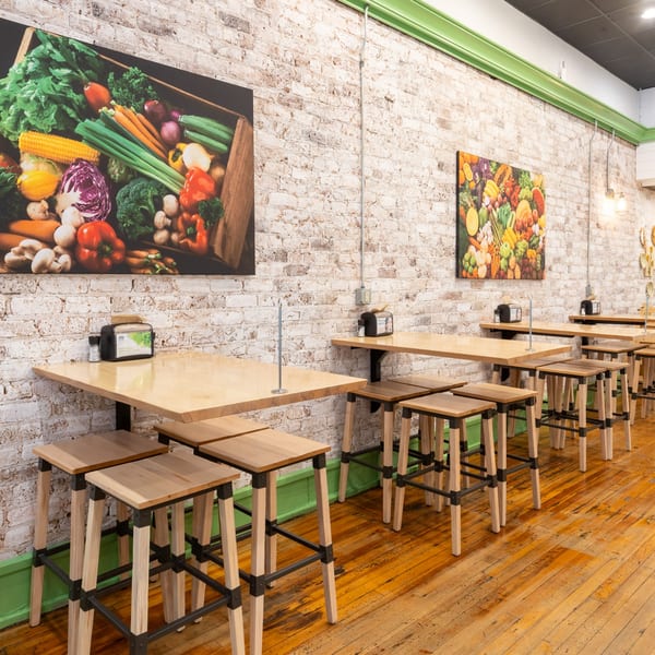 Restaurant interior with light wood tables and stools against a white brick wall with vegetable art.
