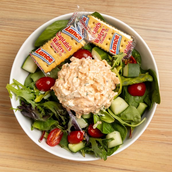 A white bowl of green salad with cherry tomatoes, cucumbers, and a scoop of chicken salad, served with two cracker packets.