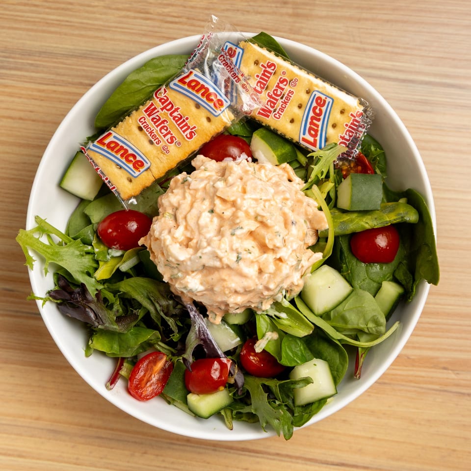 A white bowl of green salad with cherry tomatoes, cucumbers, and a scoop of chicken salad, served with two cracker packets.