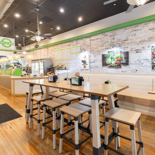 Bright, modern restaurant interior with a long communal wooden table and stools, a service counter, and brick walls.