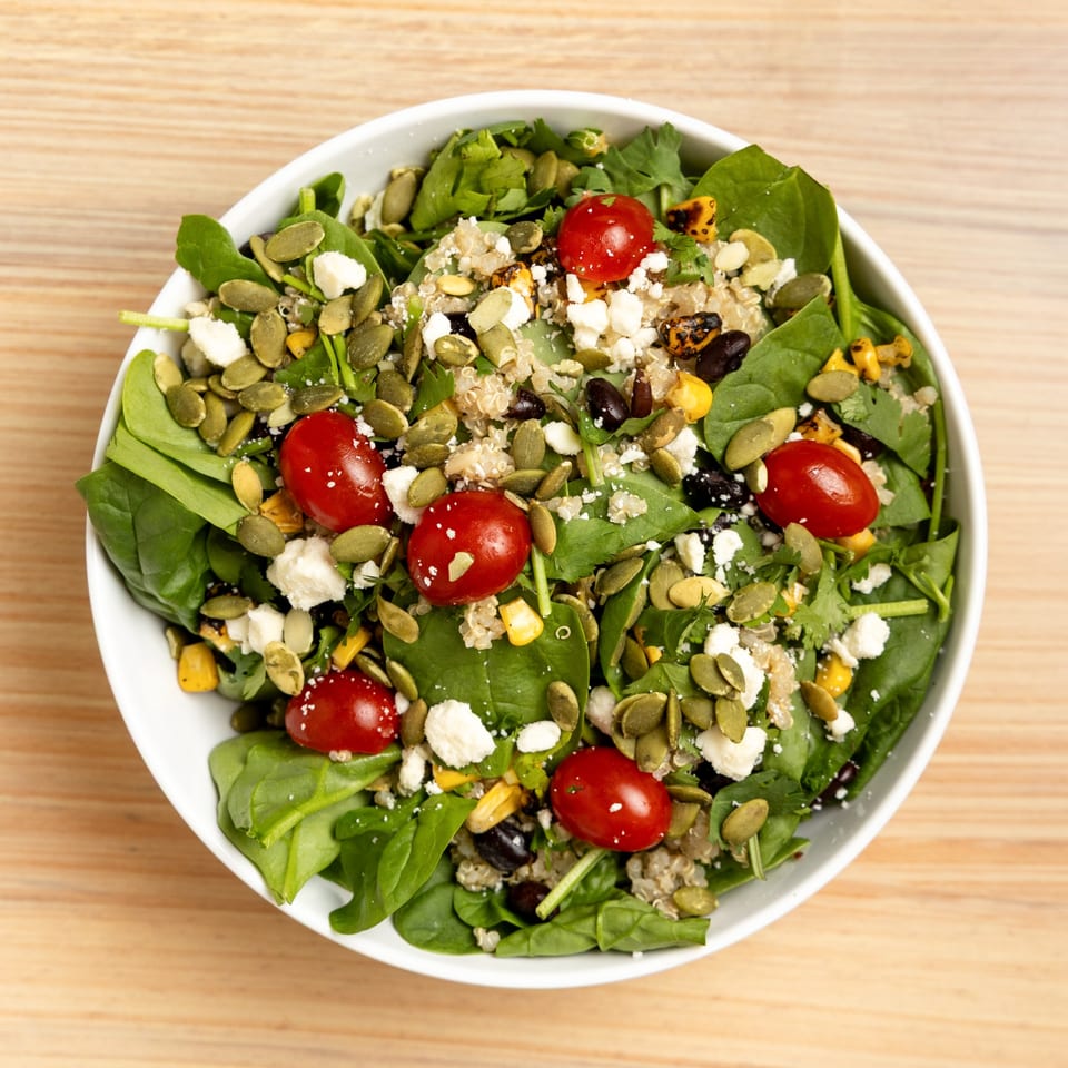Overhead view of a vibrant salad with spinach, quinoa, cherry tomatoes, feta, pumpkin seeds, and corn.